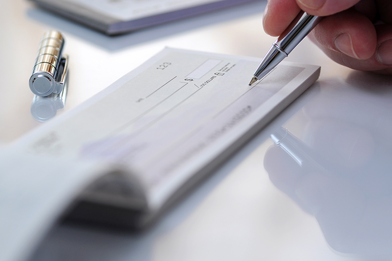 Close-up of a hand holding a pen and writing on a blank check, with a silver pen cap resting nearby on a reflective surface.