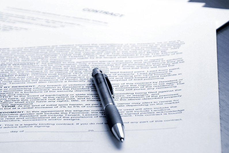Close-up of a black pen resting on top of a printed contract document, with additional sheets of paper underneath on a dark wooden surface.