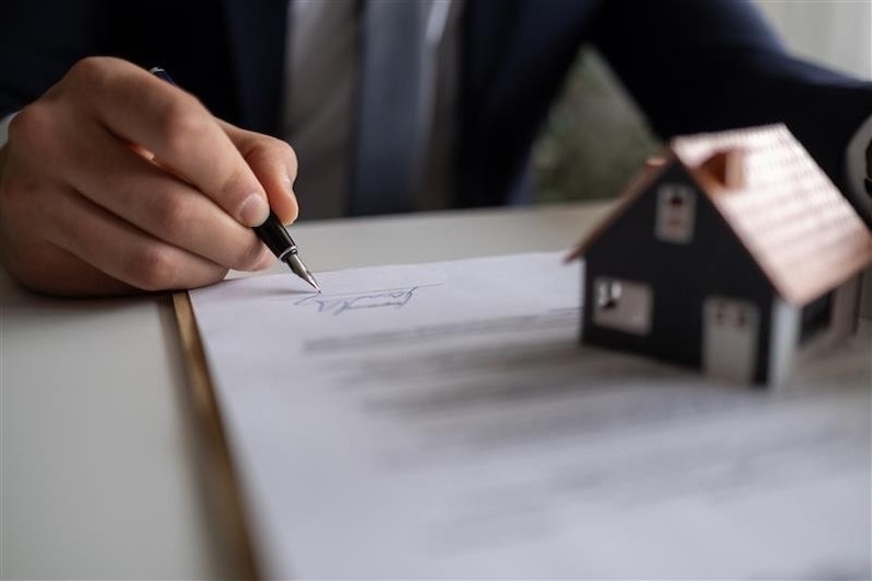 A person signing estate planning documents with a pen beside a small model house.