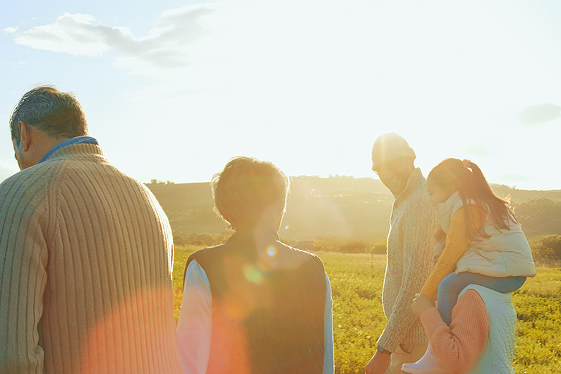 Four people walking outdoors in a sunlit field, with bright sunlight creating lens flare and a scenic landscape in the background. One person is carrying a child on their shoulders.