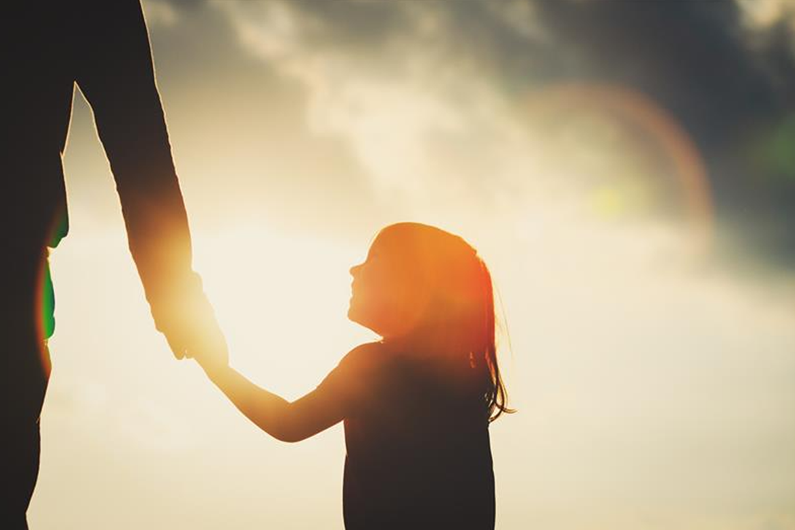 Silhouetted child holding an adult's hand against a bright, sunlit sky with clouds.