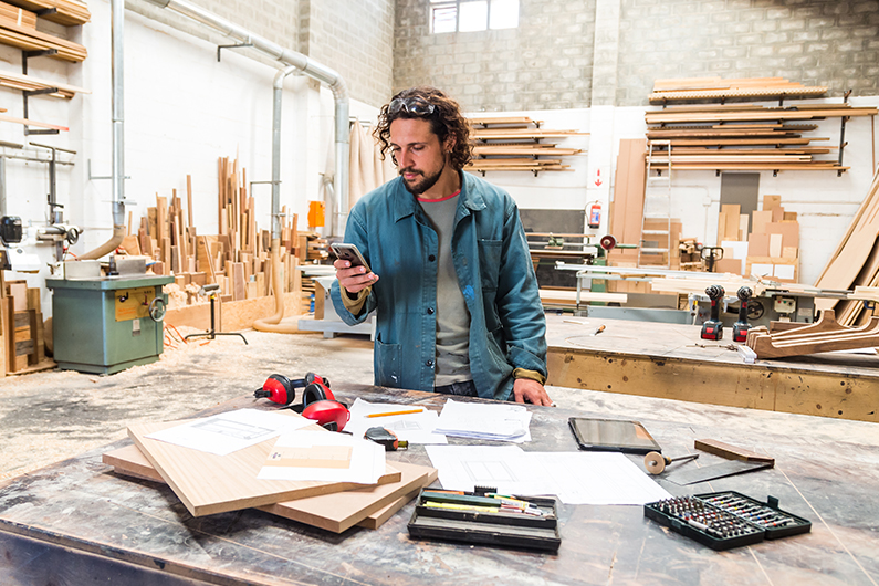 Man stands in his wood working shop and searches for small business health insurance plans on his phone.