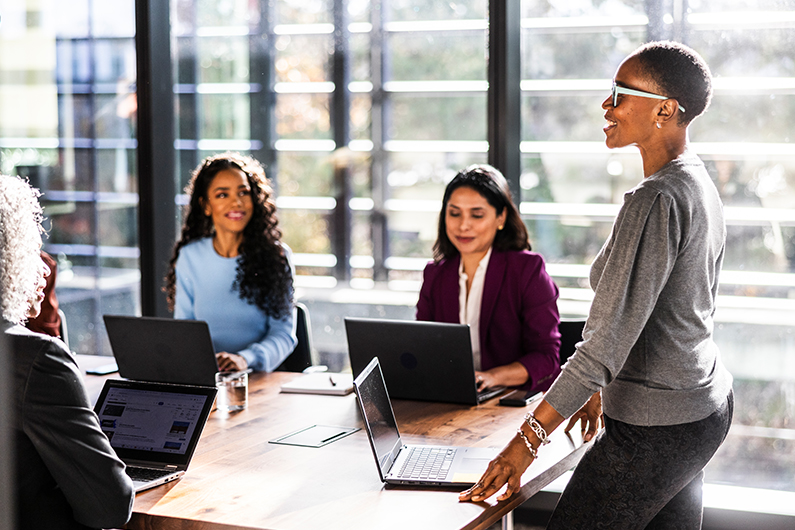 A female business owner stands while speaking to office workers in modern conference room.