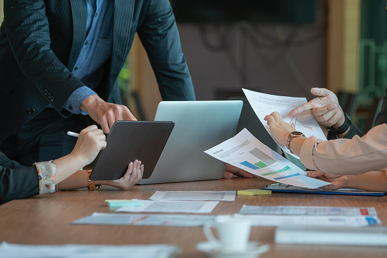Close-up of a business meeting with several people collaborating around a table. One person is pointing at a tablet, another is holding printed charts with graphs, and a laptop is open in the center. Documents and a coffee cup are on the table.