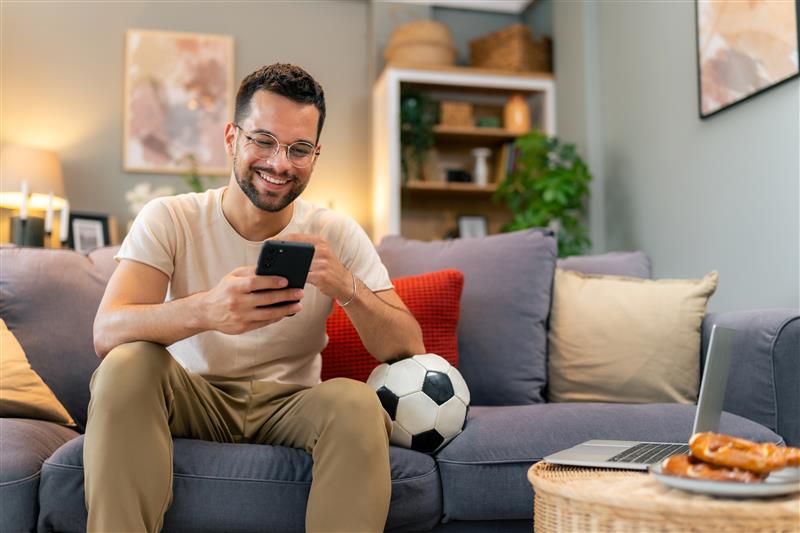 Man sits on a couch using his phone to watch a live-streamed sporting event with a soccer ball nearby.