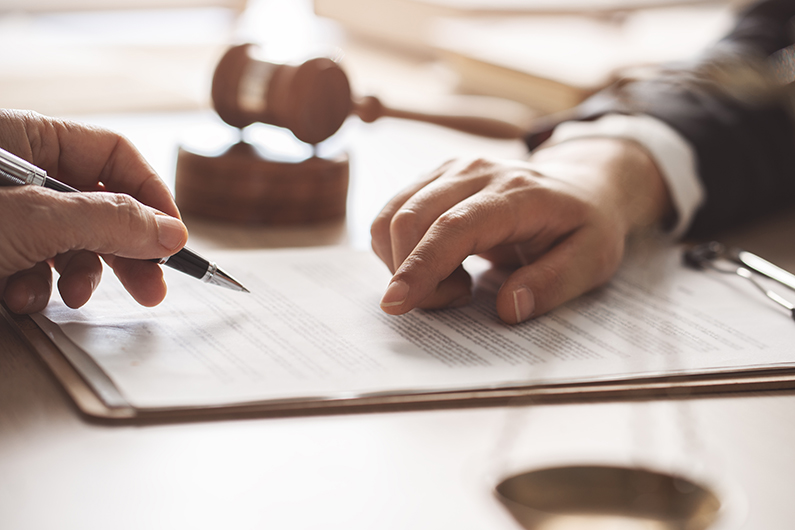 Close-up of two hands reviewing and signing a legal document on a desk, with a wooden gavel in the background.