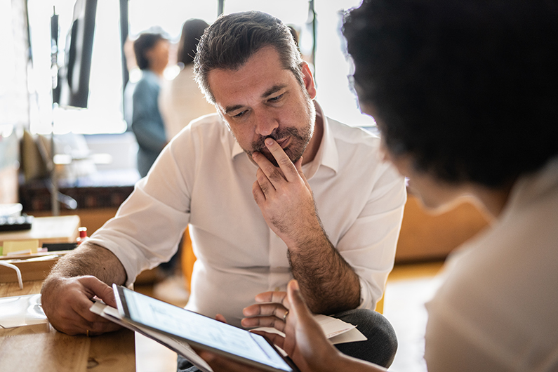 Male business owner in a white shirt sits with a woman and reviews information on a tablet screen.