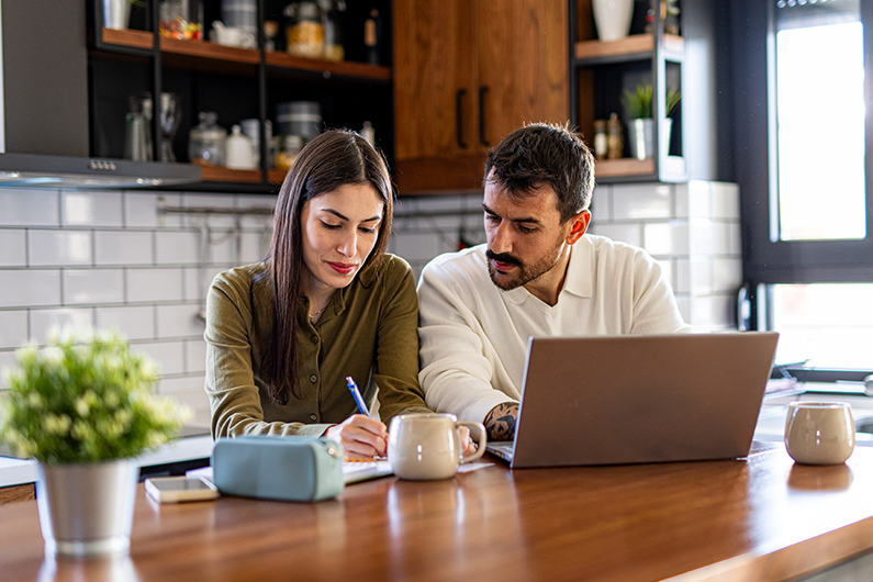 A couple sits together at a kitchen table, using a laptop to compare financial considerations of selling vs renting their home.