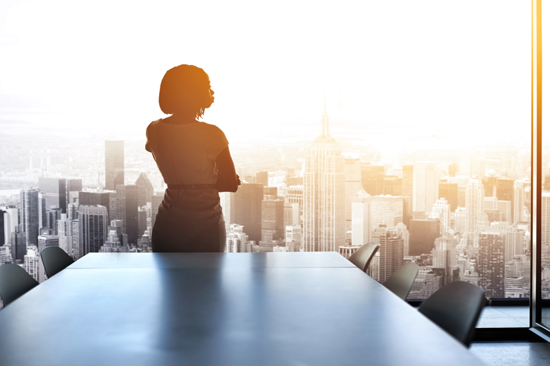 A female business owner overlooks the sky at sunset from a large office window.
