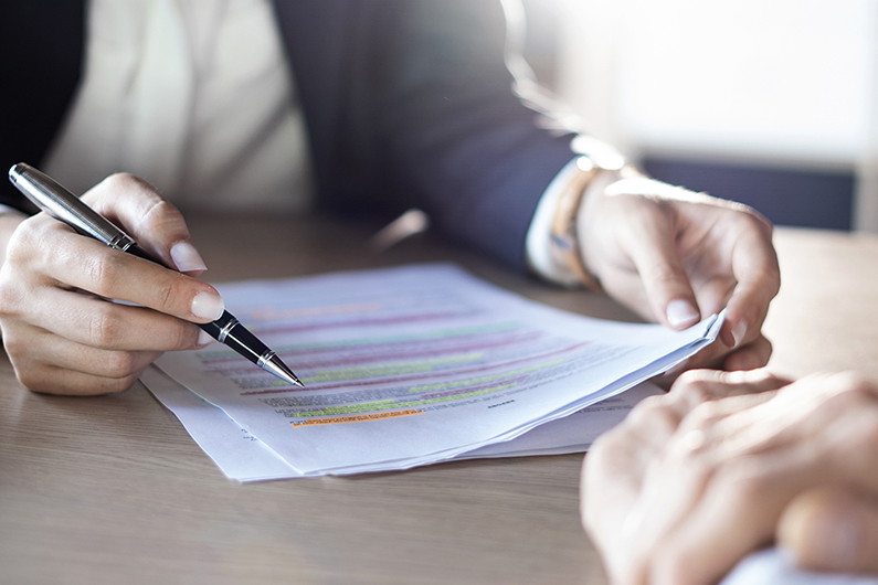 Close-up of a person holding a pen and reviewing a printed document with multiple highlighted sections in different colors on a wooden table.