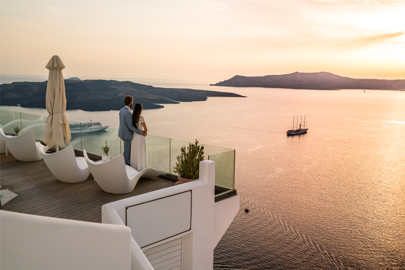 A man and woman stand on a balcony and overlook the sunset across the ocean. 