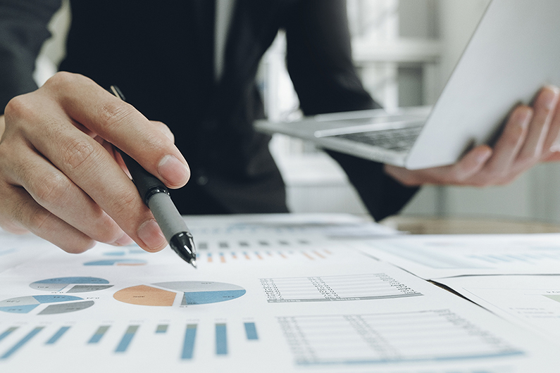 Person holding a laptop in one hand and pointing at printed charts and graphs with a pen, showing financial or business data analysis.