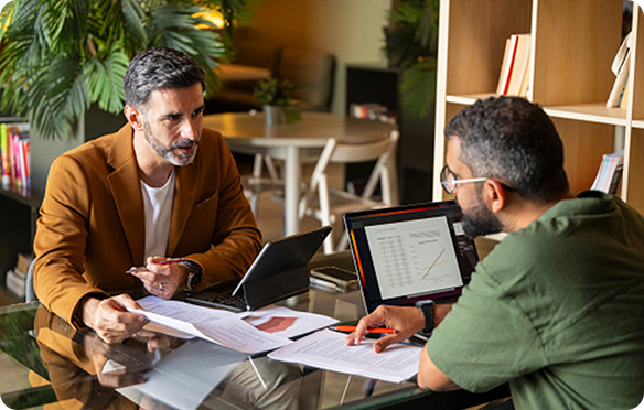 Two men sitting across form each other in front of their laptops and paperwork at a table having a discussion