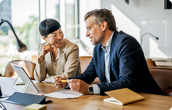 Two people sitting next to each other in front of a tablet and paperwork at a table having a discussion
