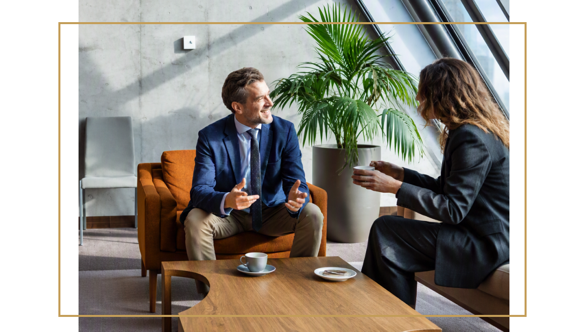 A man and woman in business attire sitting near each other at a coffee table drinking coffee and talking