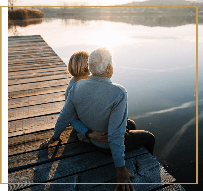 A man and woman sitting on a dock on a lake with their legs dangling over the side, looking at the sun set. 